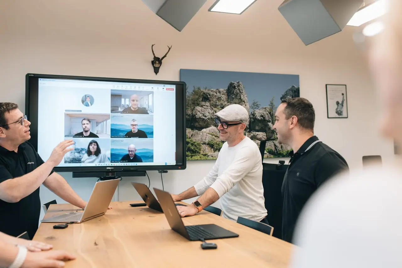 PASSION4IT workshop in a conference room: a Microsoft Teams video call with six participants is on the large screen while three people collaborate on laptops at the table.